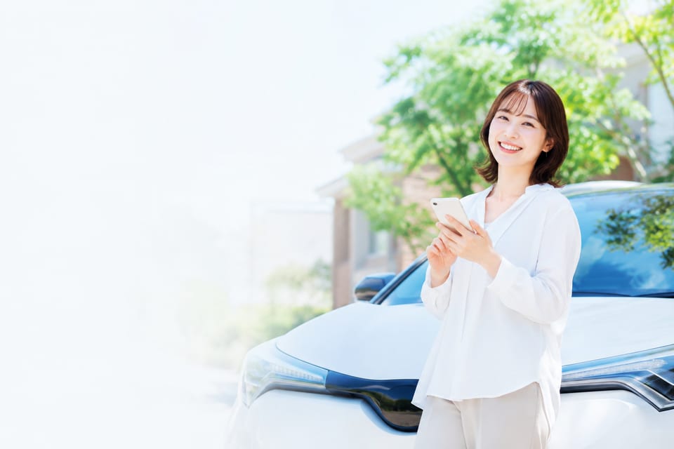スマートフォンを手に、自宅の前で愛車のとなりに立つ女性