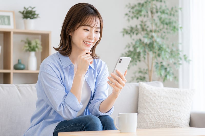 自宅のソファでスマートフォンを見て、届いた入札額を確認する女性